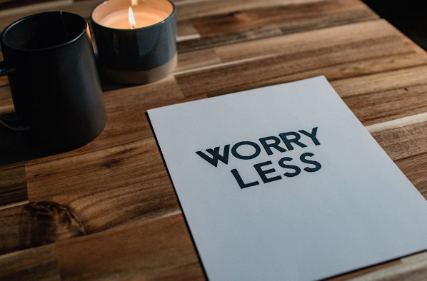 Paper that says worry Less in bold text on wooden table with two lit candles nearby. 